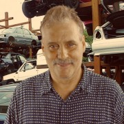 Man with light brown hair and a salt-and-pepper mustache standing in front of an auto yard